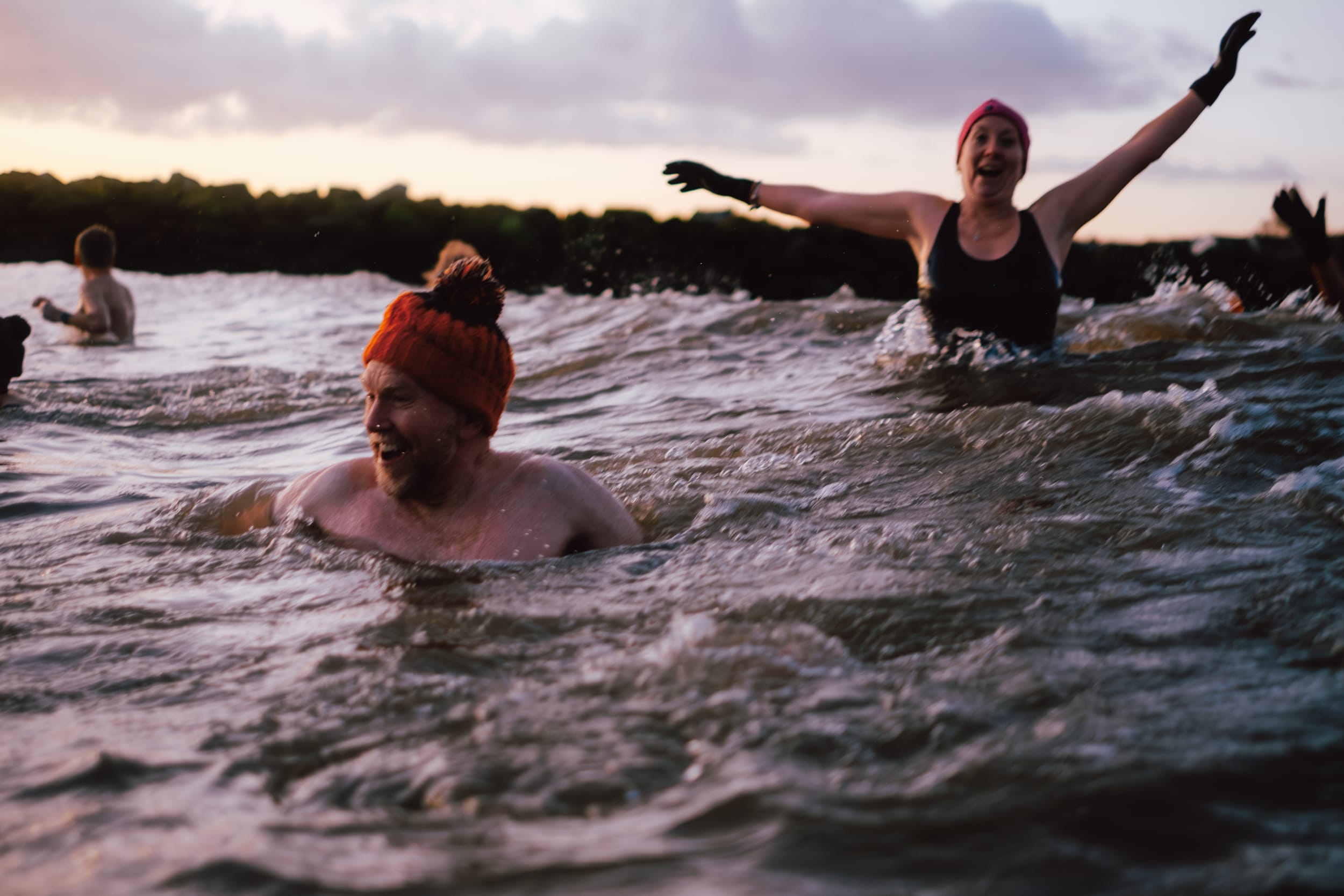 A group of people swimming in a body of water during sunset. One man with a beard is wearing an orange knitted hat and smiling while swimming. A woman in a black swimsuit, also smiling, is splashing water with her arms raised. Another person is visible in the background.