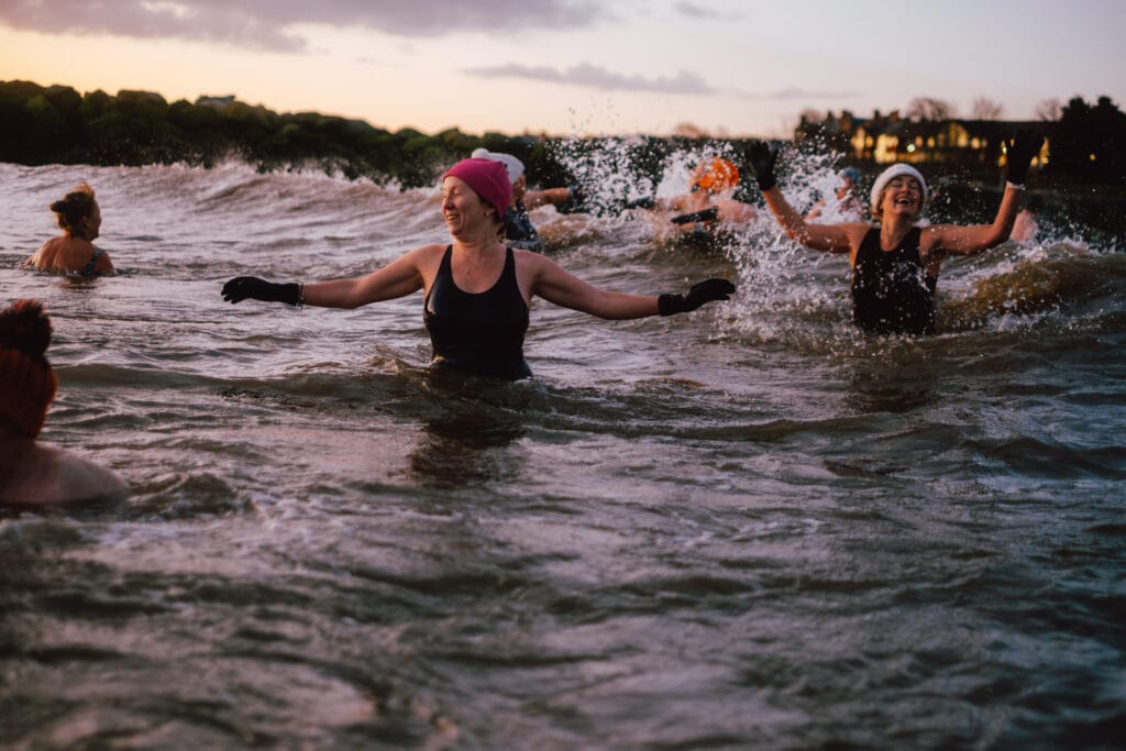 A group of women swimming in a body of water during sunset, some wearing hats and gloves. They are enjoying the water, splashing and smiling as they engage in the activity.