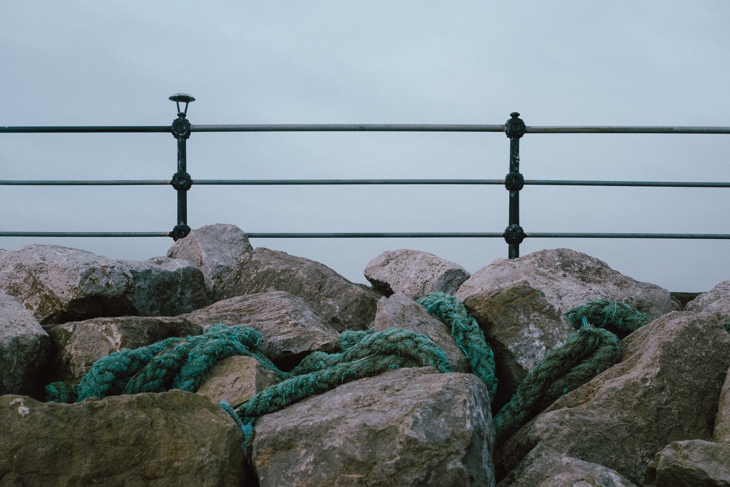 A close-up view of a rocky area with pieces of green rope scattered among large gray rocks, and a metallic railing in the background.