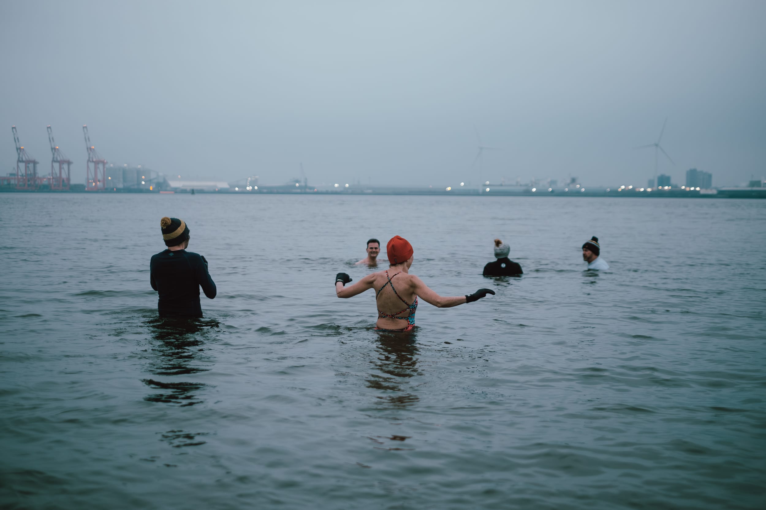 A group of people swimming in a body of water with a foggy background. One person in the foreground is wearing a swim cap and appears to be gesturing, while others are partially submerged. In the distance, there are industrial structures and wind turbines.
