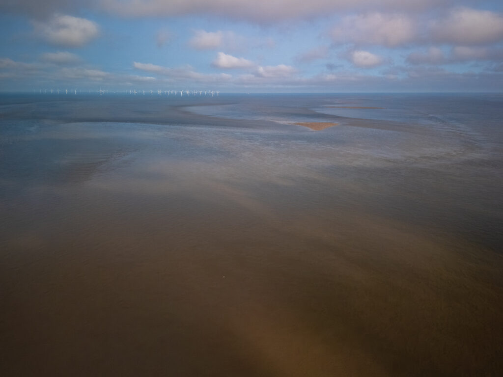 Aerial view of the River Mersey at low tide, with wind turbines in the distance and a cloudy sky.