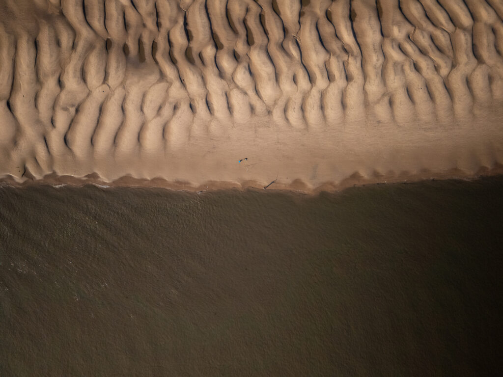 An aerial view of a beach at low tide with textured sand patterns and the dark water of the River Mersey. A person, tiny in the frame, is fishing in the centre of the photo.