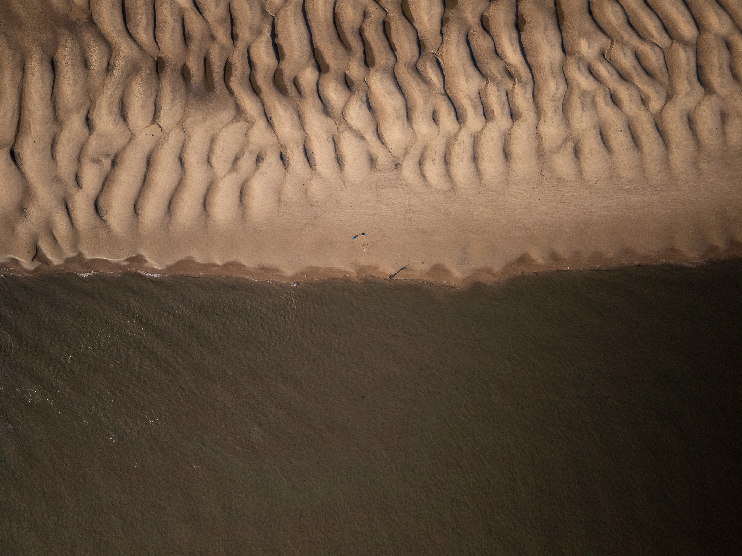 An aerial view of a beach at low tide with textured sand patterns and the dark water of the River Mersey.