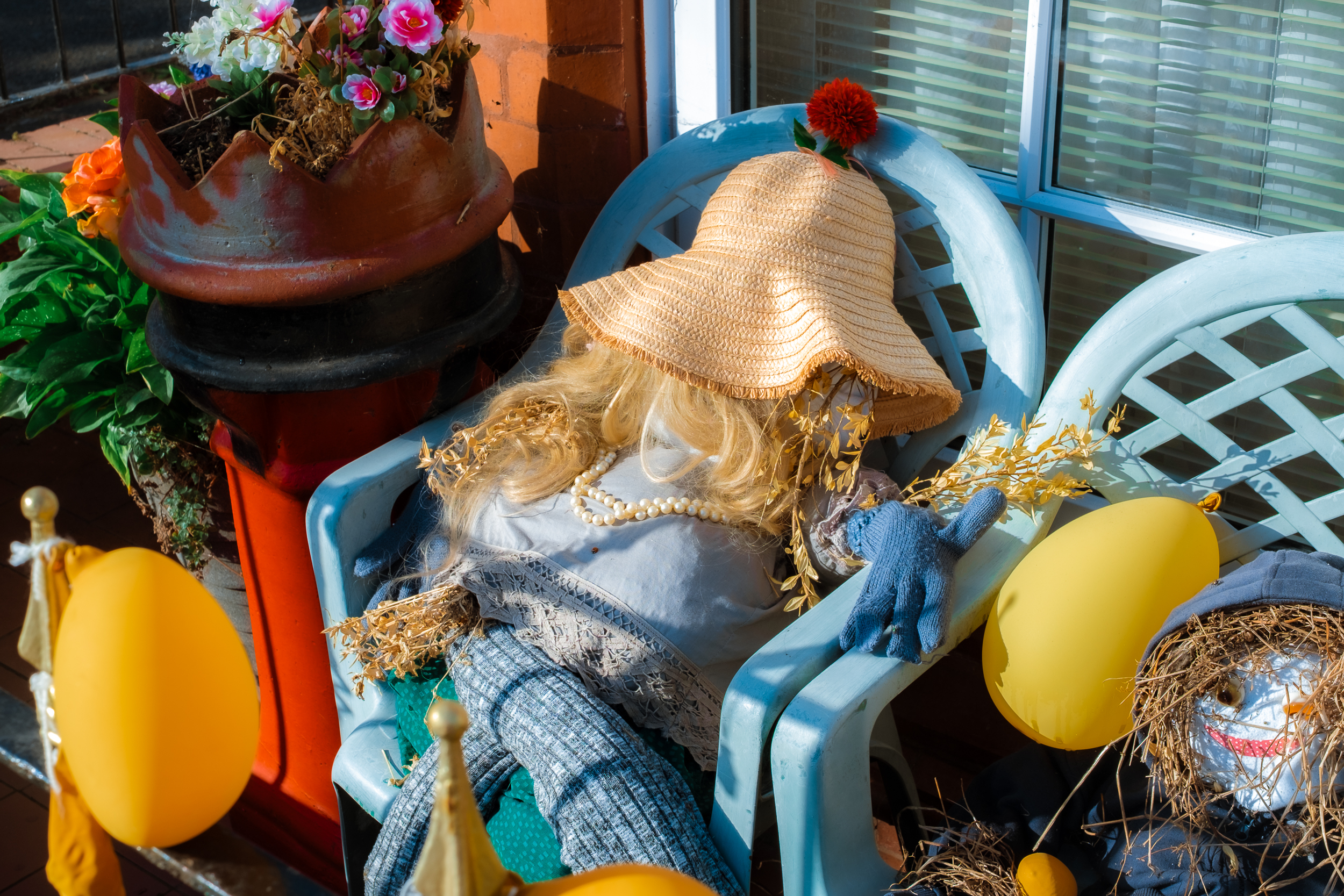A scarecrow from the New Brighton scarecrow festival wearing a straw hat and sitting in a blue plastic chair.