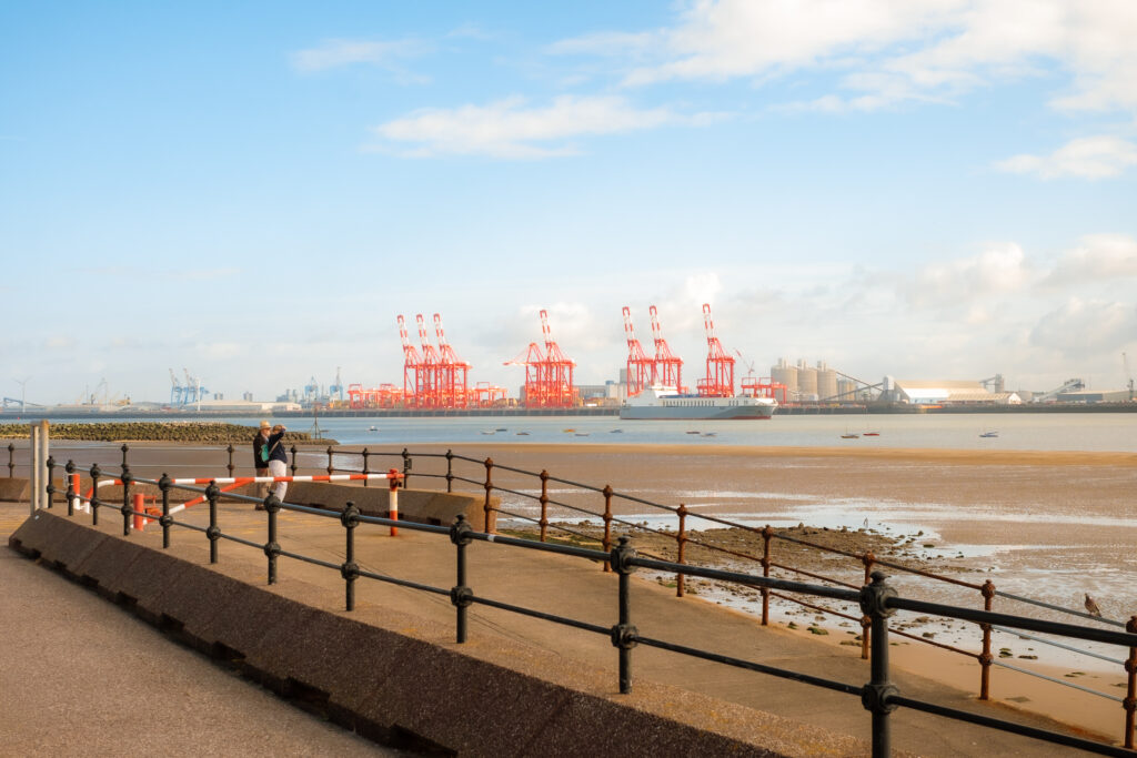 View of the Liverpool docks across the River Mersey from New Brighton, Wirral, with a person standing on a walkway.