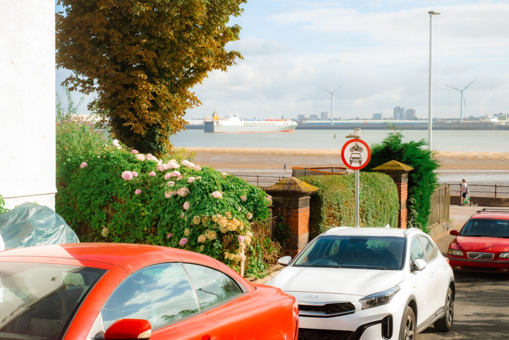 View of New Brighton, Wirral, with cars parked on the street, a large cargo ship on the River Mersey, and wind turbines in the distance.
