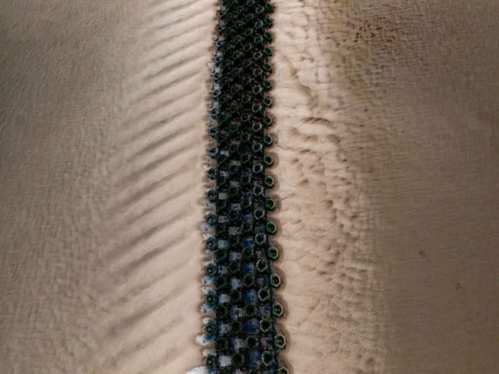 An aerial drone shot of a coastal landscape with a green and blue structure on the right side and a sandy beach on the left.