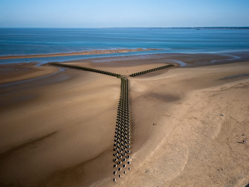An aerial view of the Y-shaped New Brighton groyne extending across a vast sandy beach towards the calm blue sea under a clear sky. The tide is out, revealing extensive sand flats with a few small figures walking near the groyne.