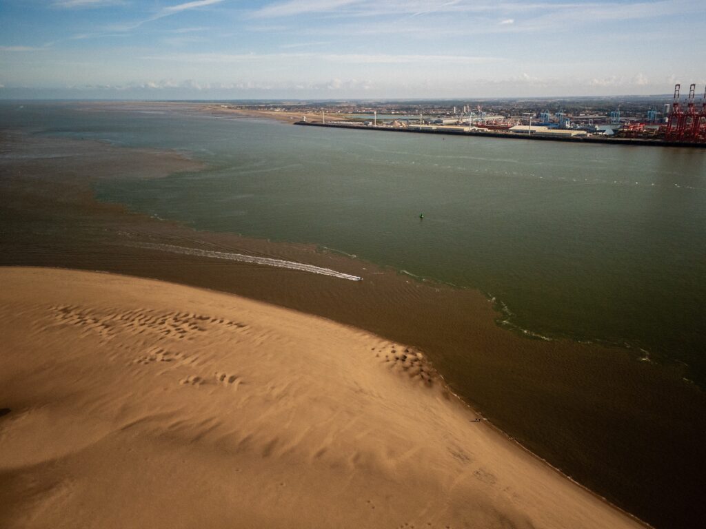 An aerial view captures a wide river mouth merging into an expansive body of water, with sandy shorelines on either side. The water exhibits varying shades of brown and green, likely due to sediment. In the distance, industrial structures and cranes are visible along the shore, contrasting with the natural landscape of the sandy beach. Wispy clouds dot the blue sky, creating a serene atmosphere.