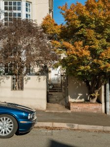 A blue car is parked on the street next to a house with a white facade. The entrance features a small pathway and an iron gate, while trees with green and orange foliage frame the scene, indicating the change of seasons. Bright sunlight casts shadows on the ground, enhancing the warm colors of the surroundings.