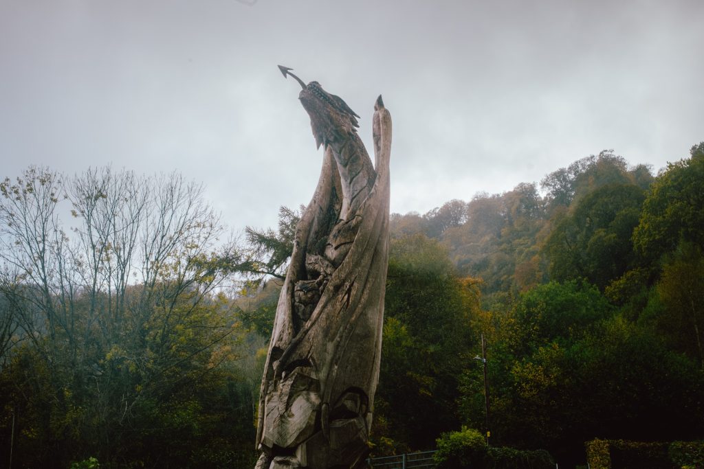 A weathered wooden sculpture stands prominently against a backdrop of misty hills and trees. The sculpture features intricate carvings resembling natural forms, with pointed elements reaching upwards. Surrounding it are various shades of green and brown foliage, indicative of autumn, while a gloomy sky adds a dramatic atmosphere to the scene.