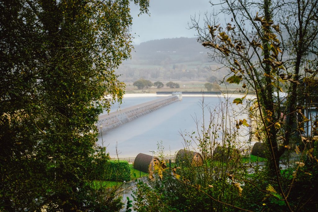 A serene landscape depicts a body of water with a long, narrow structure extending across it, possibly a dam or bridge. Surrounding the water are trees with autumn foliage, and in the background, rolling hills are shrouded in a light mist, creating a tranquil and natural atmosphere.